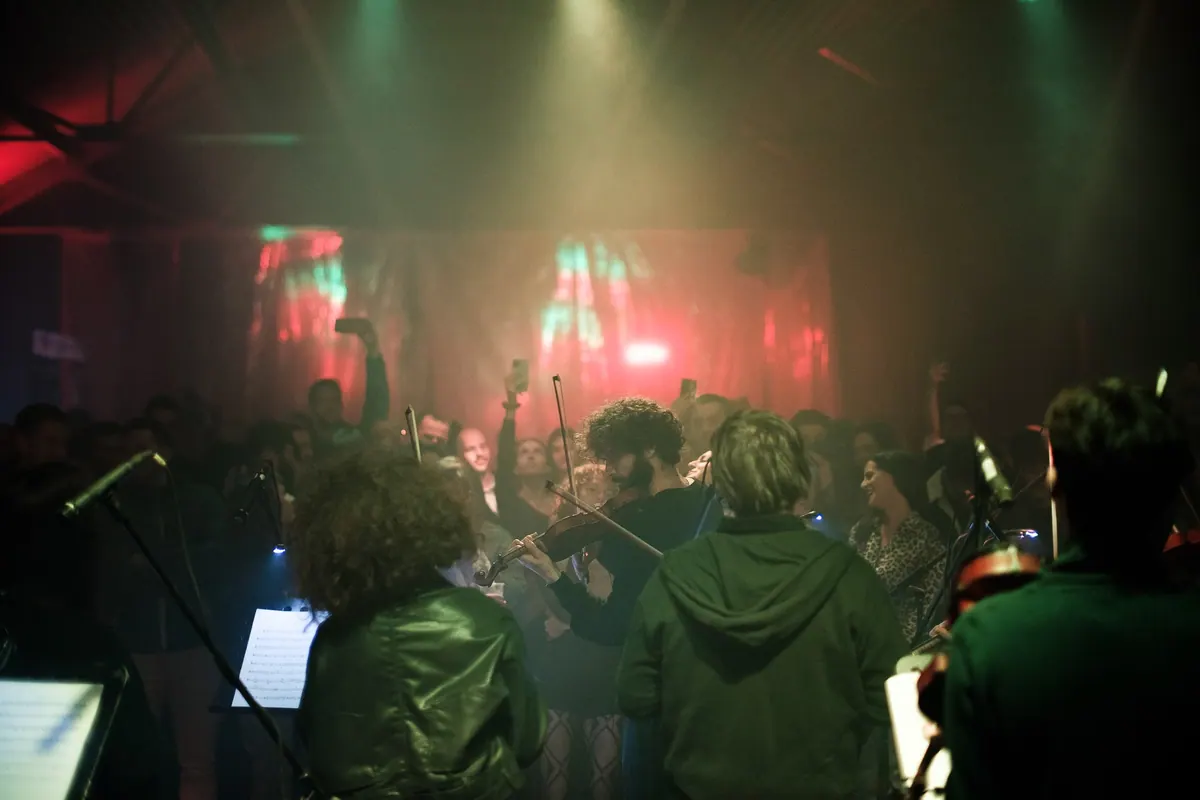 Muzičari orkestra sviraju okruženi publikom, zeleno i crveno svjetlo, ljudi snimaju telefonima