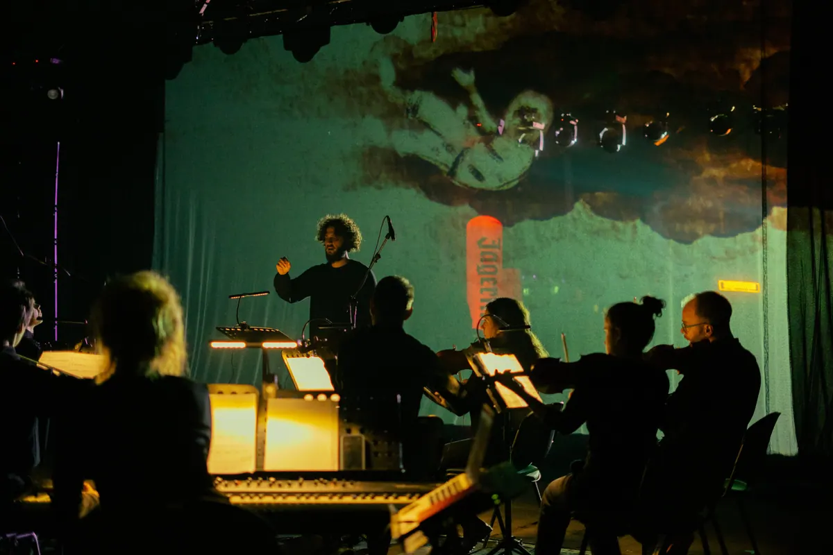 Dirigent i orkestar ispred zelene projekcije figure u svemirskom odijelu, The Stranger Within