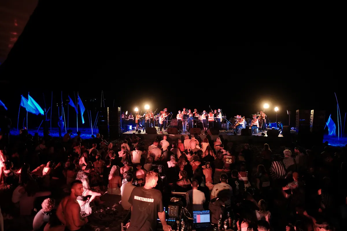 Orkestar u narandžastim prslucima na bini u daljini, plave zastave i publika u crvenom svjetlu ispred tonskog pulta